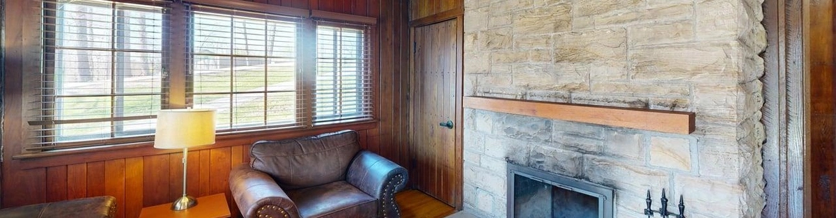 A cabin living room. On the right side of the room is a large stone fireplace with fire pokers and a wooden mantel. Next to it is a leather chair with a side table and lamp. Behind the chair are three windows.
