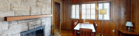 A cabin living room. On one side of the room stands a large stone fireplace complete with a wooden mantle. Next to the fireplace is a long, slender dining table with one head against the wall and lined with chairs. The walls and ceiling are wood paneled and the floor is also wooden.