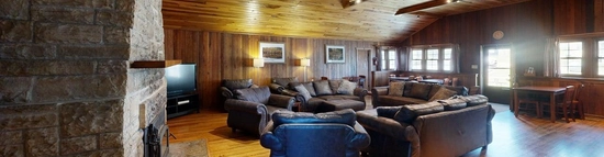 An open-concept space in a cabin. There are multiple couches and chairs in the center, right next to a stone fireplace. The walls and ceiling are wood paneled, along with wood floors. Windows are on the far side of the room, allowing some natural light into the space.