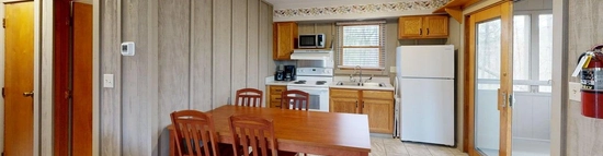 A cabin kitchen with the countertops, cabinets, and appliances lining one wall. In the foreground is a dining table with two chairs on either end. On the right side of the room is a sliding glass door. On the left side of the table, past the wall it sits against, is a hallway that extends off-screen.