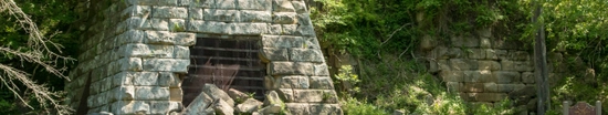 Historic iron furnace surrounded by trees and greenery in a wooded park setting. A small fenced area sits in front of the fireplace, with sunlight filtering through the forest canopy.