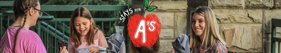 Three people sit together at an outdoor dining table on a lodge patio, smiling and enjoying time together. A hand-drawn red apple graphic with the phrase “Stay's for A’s” is overlaid in the center of the image, creating a back-to-school themed design. Stone walls and green railings frame the background.